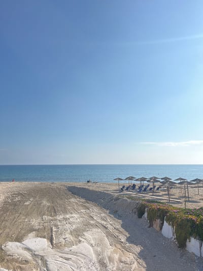 The image shows a view of a beach in Limenaria, Greece (latitude: 40.626, longitude: 24.573). The scene is captured from a slightly elevated vantage point, looking down towards the shoreline. The beach is mostly empty, with only a few people visible in the distance, sitting on lounge chairs beneath beach umbrellas. The sand is light-colored, interspersed with patches of gravel. There are several rows of lounge chairs and umbrellas lined up along the beach. In the foreground, a path leads down to the beach, with a small patch of greenery on the right side. The sky is clear and blue.