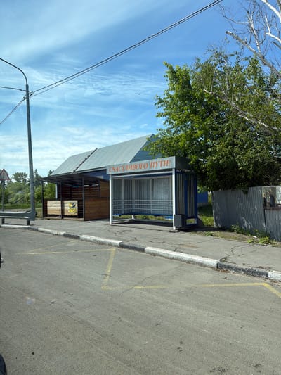 The image depicts a street scene in Aur, Russia, featuring a bus stop with a covered shelter. The shelter has a metal frame and a corrugated metal roof. The sides of the shelter are open, revealing a bench inside. Above the shelter, there is an orange sign with Russian text that reads "СЧАСТЛИВОГО ПУТИ!", meaning "Have a good trip!". To the left of the bus stop is a small wooden structure, possibly a kiosk or storage area. A metal pole with power lines runs vertically along the left side of the image. A traffic sign with three colored circles is visible further to the left. The bus stop is situated on an asphalt road with yellow markings and a concrete curb. A corrugated metal fence runs along the right side of the bus stop, and a framed image of a person in religious attire is attached to it. Lush green trees and foliage are present in the background. The sky is blue with scattered clouds.