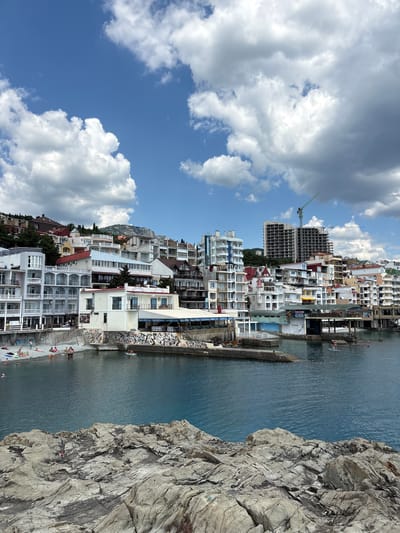 The image shows a coastal town in Kiparisnoe, Ukraine, with buildings lining the waterfront and hills in the background. The foreground features rocky terrain overlooking the blue sea. The town is densely packed with multi-story buildings of various architectural styles, many painted white with balconies and red roofs. Some buildings appear to be hotels or apartments, while others might be shops or restaurants along the waterfront. A small beach area is visible with several people sunbathing and swimming. In the water, a few individuals are engaged in activities like paddleboarding. Construction is underway on a large building in the distance, with cranes visible. The sky is blue with scattered white clouds. The overall scene depicts a lively seaside resort on a sunny day.