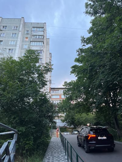 In the city of Belorechensky, Russia, a scene unfolds on a cloudy day. A multi-story, grey apartment building with visible balconies stands to the left, partially obscured by lush green trees. A walkway with grey and white patterned paving stones leads away from the viewer and up a slight incline towards another, lower apartment building visible in the background. A person in an orange outfit walks away from the viewer on the walkway. To the right of the walkway, a black SUV with illuminated taillights is driving away from the viewer on a paved road, with more trees lining the right side of the road and the image. Power lines are visible against the overcast sky.