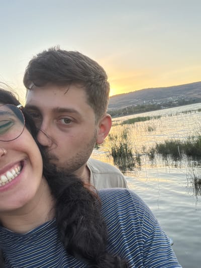 A couple takes a selfie by a body of water at sunset. The woman is in the foreground, smiling and wearing glasses and a blue and white striped shirt. The man is behind her, kissing her cheek, wearing a light-colored shirt. The background shows a body of water with reeds and a distant hillside with a town or village. The sky is a pale orange and yellow, indicating sunset. This scene is likely located in the city of Emek HaYarden Regional Council, Israel, given the landscape and vegetation.