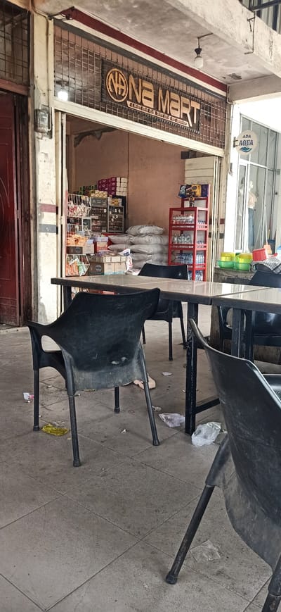 The image depicts an outdoor seating area in front of a small shop labeled "NA MART" in Tambonbaroh, Indonesia. Several black plastic chairs are arranged around dark-colored tables with reflective surfaces. The ground is paved with square tiles and littered with various pieces of trash, including plastic bags and wrappers. Behind the tables and chairs, the shop's entrance is visible, revealing shelves stocked with goods such as bottled water, packaged food items, and sacks of what appear to be rice or flour. A red shelf unit displays bottled drinks, and a sign for "ADA AQUA" is mounted on the exterior. The overall environment suggests a commercial area with an outdoor seating arrangement that appears to be in need of cleaning.