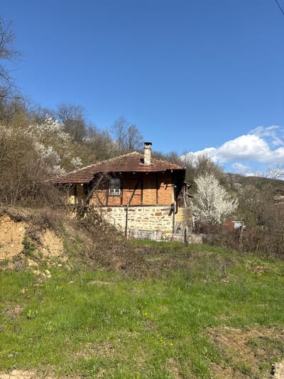 The image shows a two-story rural house situated in what appears to be the countryside. The house has a combination of brick and stone exterior walls, a red-tiled roof, and a chimney. The surrounding environment consists of grassy areas, bare trees, and blossoming white trees indicating springtime. The sky is blue with scattered clouds. The location is presumed to be Liava Reka, Bulgaria, based on the prompt. There are no people visible in the image. The house is positioned on a slight incline, with a partially obscured wooden structure visible behind it. The foreground contains dry vegetation and patches of exposed dirt.