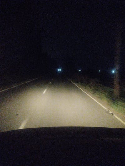 This media file depicts a nighttime view of a road, likely from the perspective of someone inside a vehicle. The primary subject is the road itself, illuminated by the headlights of the vehicle. A white dashed line runs down the center of the road, guiding the way. To the right of the road, there is a grassy shoulder with some vegetation. In the distance, several streetlights or other light sources are visible, creating bright spots against the dark sky. The overall scene is one of driving at night, possibly on a highway or a rural road. The time of day is clearly night, as evidenced by the darkness and artificial lighting. No people are visible in the frame. The image has a grainy quality, suggesting it may have been taken in low light conditions or with a lower quality camera. There is no visible text in the image. The location context provided indicates Akhali Dioknisi, Georgia, which is a geographical reference.