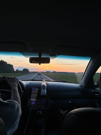 A first-person perspective shot from inside a car driving on a rural road in the city of Irnyshki, Russia, at sunset. The sun is setting on the horizon, casting a warm glow across the sky with shades of orange and yellow. The road ahead is straight and empty, flanked by green fields and trees. The car's dashboard is visible, with a phone mounted on a holder displaying a navigation app. A bottle of water is also placed on the dashboard. The steering wheel and the driver's hands are in the foreground, indicating that the car is in motion. The overall mood of the image is peaceful and serene, capturing the essence of a scenic drive during golden hour.