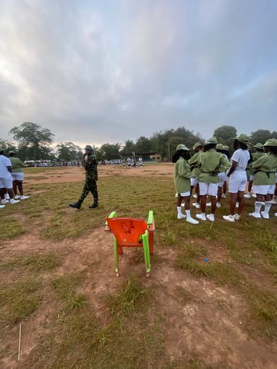 In Emure-Ekiti, Nigeria, at a parade ground, a group of young people in matching green uniforms and white shorts stand in formation. A lone figure in camouflage uniform walks across the grassy area in the foreground. In the foreground, two plastic chairs, one orange and one green, are placed on the ground. The sky above is overcast and cloudy, and trees are visible in the background. The ground is a mixture of dirt and grass, with some litter scattered around.