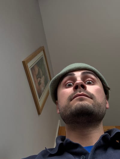 A low-angle, close-up photograph captures a male subject indoors. The individual wears an olive-green ribbed beanie hat, a dark blue collared polo shirt over a blue t-shirt, and exhibits dark facial stubble. His eyes are wide open and directed towards the camera. The background consists of light-colored walls, with a corner where they meet the ceiling. A light wood-framed picture hangs on the left wall, depicting a domestic animal with brown fur in a natural environment. The setting is an interior space, consistent with a domestic dwelling in the Cherwell District, United Kingdom. No specific address is discernible from the image.