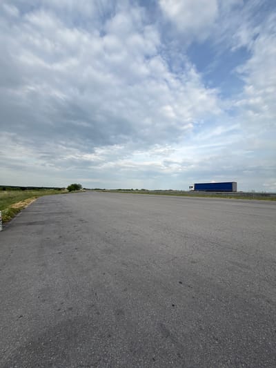 The image shows an asphalt road stretching into the distance under a cloudy sky. A large blue trailer truck is visible on the road in the distance. The roadside features a grassy area with some trees and vegetation. The location is Kolarovo, Bulgaria.
