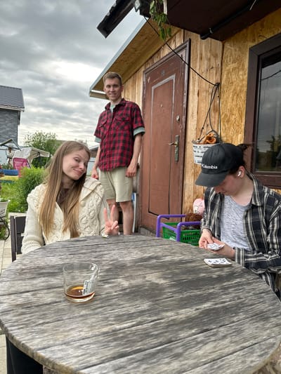 This image captures an outdoor scene featuring three young adults in a backyard setting in Bol'shaya Mos', Russia, under an overcast sky. In the foreground, a large, weathered round wooden table occupies the center of the frame. On the table, to the left, is a clear glass tumbler containing a small amount of amber liquid, with a white sticker adhered to its side. A small white daisy-like flower rests on the table's surface towards the center. Seated at the table on the left is a young woman with long, light brown hair, wearing a cream-colored chunky knit cardigan over a dark top. She is looking towards the camera with a slight smile and forming a peace sign with her right hand. To the right of the table, a person is seated, largely obscured from the chest down. This individual wears a black "Columbia" baseball cap, a grey mottled t-shirt, and a black and white plaid long-sleeved overshirt. White wireless earbuds are visible in both ears. Their gaze is directed downwards, focused on a small stack of white cards with black markings held in their hands, with another small stack resting on the table in front of them. Standing behind the table, slightly to the left of a wooden door, is a young man with short, light brown hair, wearing a red and black plaid short-sleeved shirt over a light green t-shirt, and light-colored shorts. He is smiling and looking towards the camera. The background features a rustic building, likely a private residence or dacha. The exterior is composed of vertical wooden planks on the left side of the door and Oriented Strand Board (OSB) sheets to the right. A dark brown door with the number "88" is prominent, featuring a modern handle and lock. To the right of the door is a dark-framed window. Above the door and window, a dark brown wooden soffit extends, under which a black corrugated conduit pipe is visible. A black cable runs diagonally across the wooden facade from the upper left. A white bucket-like container, with partially visible text that includes "ода", hangs from the wall near the door handle and contains brown, soft objects. Below the window, a purple plastic crate and a green plastic basket are present, alongside a brown plush toy. Further in the background, to the far left, a grey brick house with a dark tiled roof is partially visible.