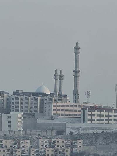 The image displays an urban landscape in Qudsaiya, Syria, under a light grey, overcast, or hazy sky. In the mid-ground, a prominent mosque features a large, white hemispherical dome and three grey minarets. One minaret is significantly taller and more ornate with multiple tiered balconies, positioned to the right of the dome. Two shorter, identical minarets with single balconies are situated further left, behind the main dome structure.

Surrounding the mosque are various multi-story buildings, predominantly appearing residential or mixed-use, constructed from light-colored concrete or stucco in shades of white, beige, and light grey. These buildings exhibit numerous rectangular windows. Communication antennas or cellular towers are visible on rooftops in the mid-ground.

The lower portion of the image depicts a densely built residential area situated on a sloping terrain. These buildings are typically 2-5 stories high, of uniform design, and constructed from light-colored materials. The underlying terrain appears dry and light-colored, suggesting arid conditions. No individuals, vehicles, or dynamic actions are discernible in the static scene.