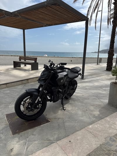 A black Yamaha MT-07 motorcycle is parked on a paved walkway near a beach. The motorcycle is angled towards the viewer and features a dark finish. In the background, the ocean is visible with a clear blue sky and scattered clouds. A wooden bench is positioned on the walkway, facing the sea. A modern, open-sided shelter with a wooden slat roof stands to the left of the motorcycle. A palm tree with fronds is visible on the right side of the frame, and a concrete planter with greenery is situated next to it. The walkway is made of concrete tiles, and a metal grate is visible in front of the motorcycle's front wheel. The scene appears to be a public area in Sesimbra, Portugal, likely along the coastline.