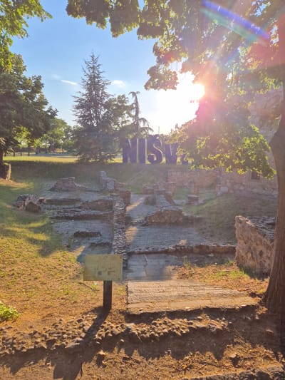 The image depicts an outdoor scene at the Mediana archaeological site in Niš, Serbia, a municipality within the larger administrative unit of Gradska opština Crveni Krst. The foreground is dominated by rough, uneven ground covered in dirt, small stones, and fallen leaves, with a paved pathway leading up towards the archaeological remains. A dark brown tree trunk is visible on the right. In the mid-ground, stone ruins of ancient structures are scattered across a grassy area. A signpost with an informational plaque stands to the left of the pathway. In the background, large, dark blue letters spelling "NIŠ" are prominently displayed against a backdrop of trees and a clear blue sky. The sun is visible through the leaves of trees in the upper right corner, casting a lens flare and bright light across the scene. The overall impression is of a historical site bathed in the warm light of the sun.