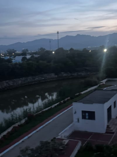 The image shows a view of Fnideq, Morocco at dusk. Mountains are visible in the distance under a cloudy sky. In the foreground, there is a building with a flat roof and dark windows, and a grassy area or small garden next to it. Streetlights are visible along a road or path near a wooded area.