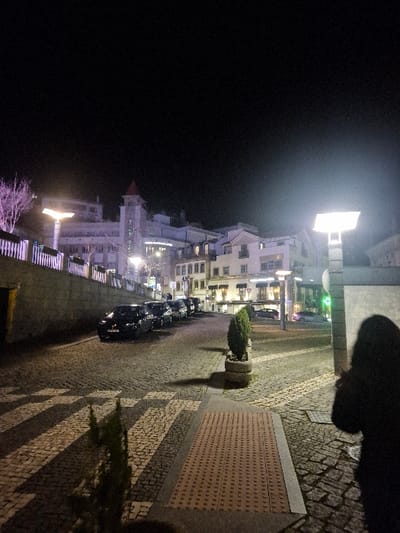 The image captures a nighttime urban street scene in Covilhã, Portugal. A cobblestone street, featuring a zebra-patterned pedestrian crossing, extends from the foreground into the mid-ground. Along the left side of the street, a line of at least five dark-colored sedans is parked diagonally. A low stone retaining wall with a white railing runs parallel to the parked vehicles. In the background, a row of multi-story buildings with traditional Portuguese architecture is visible; one prominent building features a red-roofed tower. Several windows in these buildings are illuminated. Overhead, the sky is dark. Artificial illumination is provided by streetlights, including one on the far left casting a purple light on a leafless tree, and another bright, square-headed streetlight prominently positioned on the right foreground. On the foreground sidewalk, a rectangular section of red tactile paving is present. A dark, indistinct human silhouette, appearing to wear a hooded garment, is partially visible on the far right edge of the frame.