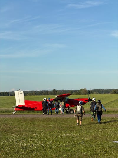 An exterior, eye-level, vertical, medium shot captures a bright, sunny day at what appears to be a small airfield. The sky is a clear, light blue with thin, wispy clouds. In the foreground, a lush green field is dotted with small white and yellow wildflowers. A paved surface runs through the middle ground, bordering the field and serving as a runway or taxiway.

Dominating the mid-ground is a red and white propeller plane, possibly a Pilatus PC-6 Porter, parked on the paved surface. The plane's tail is prominent, with "YL-LZI" written in black text on its fuselage. Several people, dressed in skydiving gear including helmets, jumpsuits, and backpacks, are gathered around the aircraft. Some appear to be boarding or disembarking, while others are walking away from the plane towards the right of the frame.

The individuals are wearing various colored jumpsuits, including blue, black, and camouflage patterns, with brightly colored helmets and backpacks. The atmosphere suggests preparation for or the conclusion of skydiving activities. In the background, a dense line of green trees forms the horizon, separating the airfield from the sky. The overall scene is one of outdoor recreation and aviation. No specific landmarks within the city of Limbaži, Latvia, are identifiable.