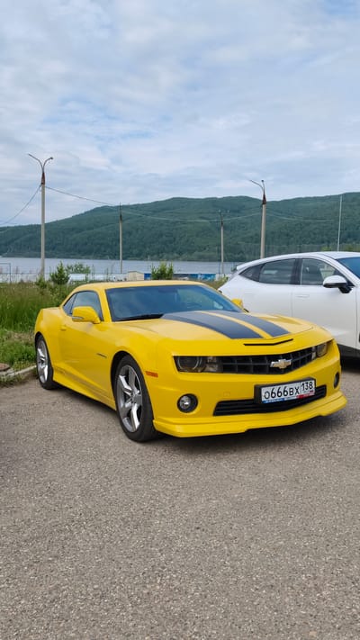 A bright yellow Chevrolet Camaro is parked on an asphalt surface in front of a body of water and a tree-covered hillside under a cloudy sky. The car has black racing stripes on its hood and a Russian license plate that reads "0666BX 138". To the right of the Camaro, a white SUV is partially visible. In the background, there are several utility poles and a section of a fence. The scene appears to be a scenic overlook or park area.