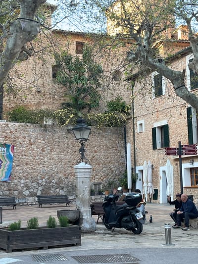The image depicts a daytime scene in a public square or plaza in Fornalutx, Spain. The square features a paved ground surface and is surrounded by multi-story buildings constructed from rough-hewn, light brown stone. These buildings possess multiple windows, many with dark green wooden shutters, some open and some closed.
In the center-left, a prominent dark metal street lamp with ornate detailing rests atop a light grey stone pedestal, which also incorporates an old, weathered stone basin, suggesting a fountain. To the left of this structure, a lower stone wall with yellow-flowering bushes on top supports a higher stone building. A colorful banner with the text "BM Internacional de les Dones" (International Women's Day) is affixed to this lower stone wall.
Multiple mature trees with light grey, mottled bark and sparse foliage due to the season are scattered throughout the scene, framing the view. Several dark metal benches are positioned around the square. In the foreground, a dark wooden planter box contains small green plants.
A black scooter with a black top case is parked facing right, to the right of the lamppost, displaying the license plate "0581 MLK." Further right, two individuals are seated on a stone ledge or bench against one of the stone buildings. One individual, a bald man, wears a dark top and dark trousers and appears to be looking down at an object in his hands. Another individual with dark hair, wearing a dark top, is seated beside him, seemingly engaged in interaction. Behind them, several white, folded outdoor umbrellas lean against the building.
A black signpost with multiple directional arrows pointing to local amenities and destinations (e.g., "Ajuntament," "Zona D'aparcament," "Policia Local") is visible on the far right. The sky is visible above the buildings and trees, indicating clear conditions. Two retractable silver bollards are positioned in the paved area on the right.