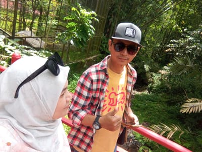 In Durin Serugun, Indonesia, two individuals, a woman and a man, are pictured from the chest up, positioned near a red railing. The woman, on the left, wears a light-colored hijab adorned with a subtle floral pattern and a pair of black sunglasses resting on top. Her profile is visible as she looks to the right. The man, on the right, wears a grey baseball cap with a patch, dark sunglasses, a yellow t-shirt with a red graphic, and a red and black plaid long-sleeved shirt unbuttoned over it. He is giving a thumbs-up gesture with his right hand and holding a smartphone in his left. The background reveals a lush, green environment with bamboo trees and dense foliage, suggesting a park or a natural setting. A cage-like structure is visible in the left background. The lighting indicates it is daytime.