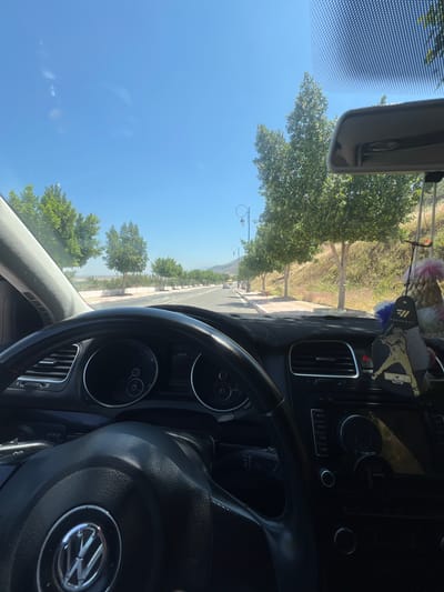 The image, taken from inside a vehicle, presents a view of a road in Beni Mellal, Morocco. The dashboard and steering wheel of a Volkswagen are prominent in the foreground, partially obscuring the view. The road stretches out into the distance, lined with trees. Street lamps are visible along the road. The sky is a clear blue, and there appears to be water in the background to the left. Other vehicles are visible on the road ahead.