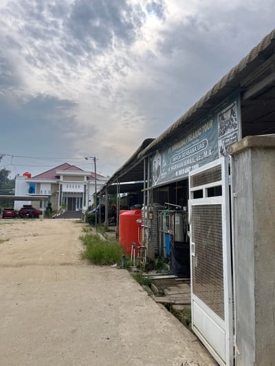 The image shows an outdoor scene in Tanahterban, Indonesia, with a modern white building with a red roof on the left, and a covered outdoor area with industrial equipment on the right. A dusty dirt road leads towards the building, with two red cars and one black SUV parked in front of it. The building features a grand entrance with stairs and decorative columns. To the right, a green banner advertising "PT. NAWADAH ISLAMIC TOUR" is visible, along with images and contact information. Industrial tanks, pipes, and other machinery are present under the covered area, with a large red water tank prominently displayed. Overcast skies with visible clouds dominate the upper portion of the image. The overall environment appears to be a mix of residential and commercial, with a slightly unkempt appearance in the foreground.