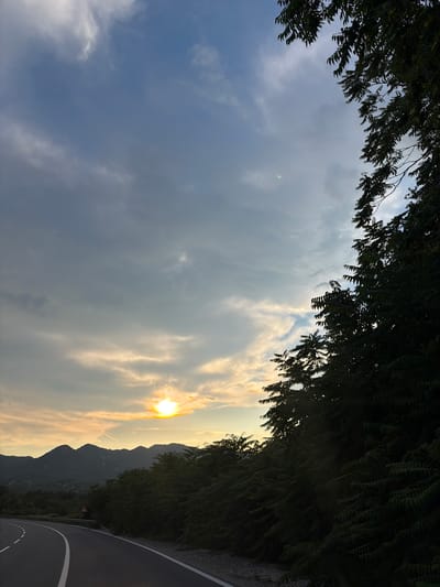 This media file captures a scenic view of a winding road bordered by lush green foliage on one side and a mountainous landscape on the other. The sun is setting, casting a warm glow across the sky and illuminating the clouds. The time of day is late afternoon or early evening. The weather appears to be clear with some scattered clouds, creating a dramatic sunset effect. The location is identified as Gospoština, Montenegro, and the presence of a road with white lines and a triangular warning sign suggests it's a public road, possibly in a mountainous or rural area. The overall mood is serene and picturesque, evoking a sense of peace and natural beauty. There are no people or specific objects of interest besides the road, foliage, mountains, and the sunset. The image is oriented vertically.