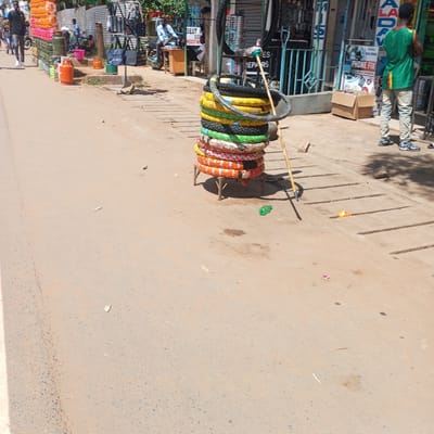 The image captures a street scene in Sheywe ward, Kenya. A stack of tires, various colors and patterns, sits on a small, makeshift wooden stand on the sidewalk. A long, wooden pole leans against the stack. Behind the tires, shops are visible, some displaying signs like "PHONE FIX" and "BEST REPAIR MEDSTOWN." A person in a green and yellow shirt stands near the shops. Other individuals are seen walking or sitting near storefronts in the background. Gas cylinders are stacked against a building in the midground. The road is cracked and shows signs of wear. The overall environment suggests a busy, commercial area.