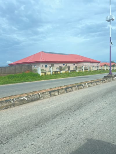 This image shows a street scene in Kona Garu, Nigeria. The foreground features a paved road with a concrete curb on the left. To the left of the curb, a strip of green grass separates the road from a building. The building has a red roof and appears to be a commercial or residential complex with multiple windows and a fence surrounding it. Further back, more buildings with red roofs are visible, suggesting a developed area.
In the upper left portion of the image, a purple and white pole with some white boxes attached to it is visible against a pale blue sky with scattered clouds. The overall impression is of a bright, possibly sunny day, although the clouds suggest it might be partly cloudy. The scene appears to be set during daylight hours. No people are visible in the image, and there are no discernible emotions conveyed. The image is taken from a slightly elevated perspective, possibly from a vehicle. There is no visible text in the image.