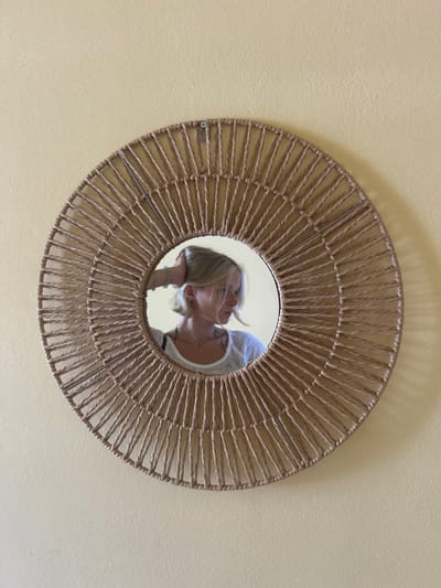 This is a medium shot, taken from a high angle, of a circular mirror with a woven frame. The mirror is hanging on a pale yellow wall. The mirror reflects two individuals. One person, with blonde hair, is leaning in towards another person, whose face is partially obscured by the first person's head. The individual with blonde hair is wearing a white top and has a bracelet on their wrist. The overall mood appears intimate and affectionate, suggesting a moment of closeness between the two people. The lighting suggests daytime, and the setting seems to be indoors. There is no visible text in the image.