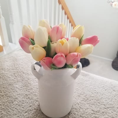 The image shows a close-up, angled view of a bouquet of tulips in a white jug. The tulips are a mix of soft pink and cream colors, with some green leaves visible. The jug has two handles and appears to be made of metal with some distressed detailing, like black marks near the handles. The arrangement is placed on a textured, light-colored surface, possibly a carpet or rug.
In the background, a wooden railing and white banisters of a staircase are visible. The lighting suggests it is daytime, and there are no indications of specific weather conditions. The overall scene evokes a sense of simple, domestic decoration. There is no visible text in the image. The context provided places the location in Virginia Beach, United States of America.