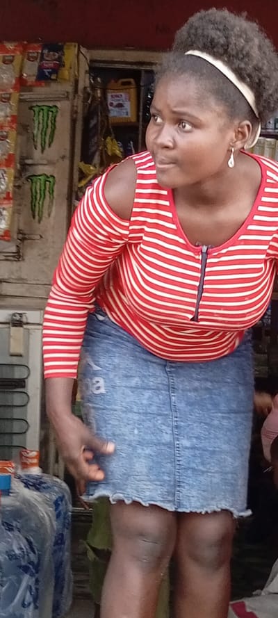 A woman with dark skin, wearing a red and white striped, cold-shoulder long sleeve shirt and a denim skirt, is partially visible. She wears a white headband in her afro-textured hair and a dangle earring. A shop-like setting is in the background, with stacks of products visible on shelves. A cooler and a water jug are also visible. The woman looks to the right.
