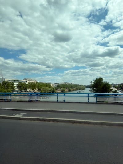 The image captures a view from a bridge overlooking a river and a city skyline. The sky is filled with dramatic, billowy white clouds against a bright blue sky, suggesting a pleasant, possibly midday, weather. A blue metal railing with a diamond-shaped mesh pattern runs along the edge of the bridge.
Below the bridge, a calm river flows, with a small boat visible on its surface. The riverbank is lined with trees and buildings, forming a picturesque urban landscape. The architecture appears to be a mix of modern and older styles. The foliage is lush and green, indicating it's likely springtime or summer.
The right side of the image shows a paved road, presumably the bridge's roadway, with white markings indicating lanes and a bicycle symbol. The road surface appears to be asphalt. The overall scene evokes a sense of urban tranquility, with nature and city life coexisting. There are no people clearly visible in the image, and the focus is on the landscape and urban elements. The time of day seems to be daytime, given the bright sky and lighting.