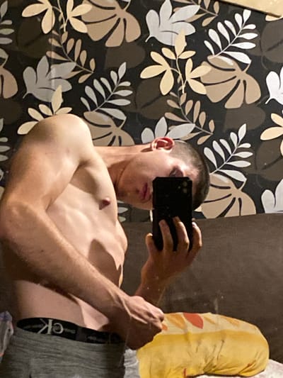 The image is a selfie taken by a person indoors, possibly in a bedroom. The background features wallpaper with a floral or leaf pattern in shades of brown, beige, and white. The person, appearing to be a young male with short hair, is shirtless and wearing grey underwear with a Calvin Klein logo band. He is holding a black smartphone to take the photo, and his face is partially obscured by the device as he looks into the camera. To the right of the person, there is a yellow object with a pattern resembling a baguette or loaf of bread. The overall lighting suggests an indoor setting, possibly with artificial light. The image does not contain any identifiable landmarks or features that would suggest a specific location within Gorna Malina, Bulgaria.