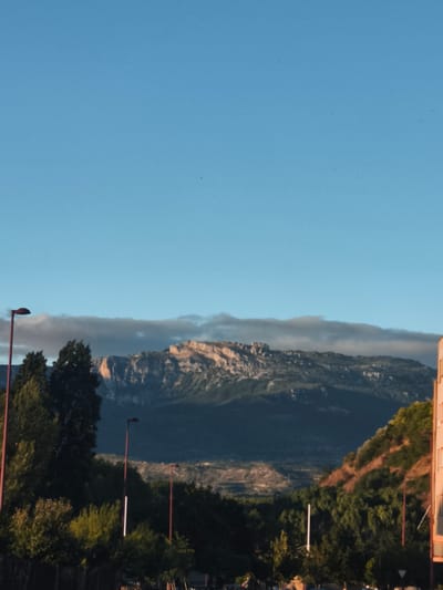 A vertical, eye-level, outdoor shot shows a mountainous landscape in the distance, under a clear blue sky with a few faint, wispy clouds. In the foreground, there are trees and two streetlights with red poles. A building with an orange facade is visible on the right side of the frame. The image appears to be taken in the city of Haro, Spain.