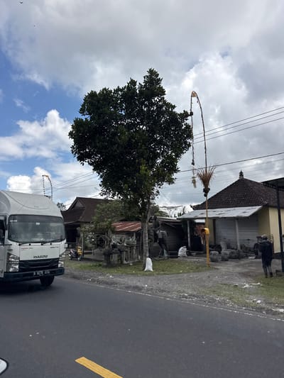A white Isuzu truck, licensed B 9278 UYA, is parked on the side of a wet asphalt road. A yellow line is visible on the road surface, suggesting it may be a lane marker. To the left of the truck is a grassy area with some debris. Beyond the grass, a building with a tiled roof and a light-colored facade is visible. In front of the building, there are stacks of stone objects, possibly decorative or for construction. A tall, thin bamboo pole with some decorative elements at the top, including what appears to be dried grass, is erected near the building and extends upwards towards power lines. A large tree with dark green foliage is situated behind the bamboo pole. The sky is cloudy, with patches of blue sky visible, indicating it is daytime. The overall scene appears to be a roadside in a semi-rural or commercial area in Susut, Indonesia. There are no people prominently featured in the scene, but a person can be seen in the background near the building. The atmosphere is neutral, with no clear indications of a specific event or emotion.