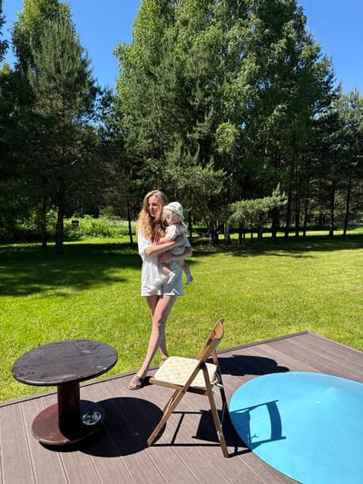The image captures an outdoor scene in Копнино, Russia, on a bright, sunny day. An adult female, with long blonde hair and wearing a white t-shirt and shorts, stands on a vibrant green lawn. She is holding a baby, who is dressed in a light-colored bucket hat, a striped top, and shorts. Both individuals are looking towards the right side of the frame. In the immediate foreground, a dark brown wooden deck is visible, featuring a round wooden table with an ashtray on its lower tier, a wooden folding chair with a light-colored cushion, and a large, partially visible blue oval object. The background consists of a dense treeline composed of tall pine trees and deciduous trees with light bark, indicative of birches, bordering the expansive green lawn. A clear blue sky is visible above the trees, and strong sunlight casts pronounced shadows from objects on the deck.