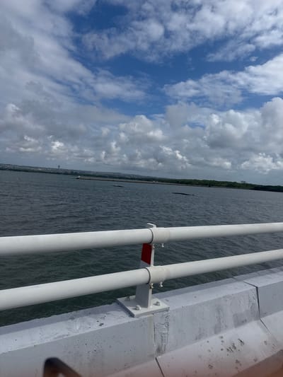 The image depicts a view of the sea and a distant coastline from what appears to be a bridge. In the foreground, a white metal railing runs horizontally across the frame, supported by a grey concrete structure. The sea is dark and choppy, with visible waves. On the horizon, a green, treed coastline is visible, with some buildings and a tall, slender structure that might be a tower or a communication mast. The sky is partly cloudy with large white cumulus clouds against a bright blue sky. The overall impression is a coastal scene on a partly cloudy day. The location is identified as South Denpasar, Indonesia, suggesting this is a view from a bridge in or near the city, overlooking the ocean.