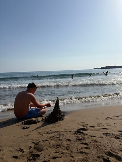 In Vlichada, Greece, a young male wearing blue swim trunks and a baseball cap sits on a sandy beach facing the ocean. He is building a sandcastle, his hands positioned near the top of the structure. The ocean stretches out before him with gentle waves reaching the shore. Several other people are visible in the water, some swimming and others wading. The sky is clear and blue, indicating a sunny day. The beach has scattered footprints in the sand, and in the distance, a rocky coastline is visible.