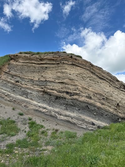 The image shows a geological formation composed of layered volcanic rock. The layers vary in color from light brown to dark gray and appear to be tilted. The formation is partially covered by green grass and some small plants, suggesting an outdoor, natural environment. The sky above is blue with scattered white clouds. The scene appears to be a natural landscape rather than an urban setting.