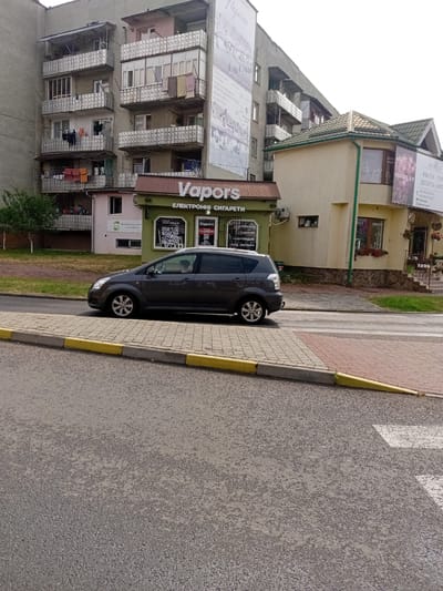 This is a street-level, eye-level, medium shot taken on a cloudy day, possibly in the late morning or early afternoon, in Tiachiv, Ukraine. The scene is dominated by buildings and a street with a car. On the left, a multi-story apartment building with many balconies, some with laundry hanging, takes up a significant portion of the frame. Below the apartment building, a small, light pink structure is partially visible.
To the right of the apartment building, a green storefront with the prominent text "Vapors" and "ЕЛЕКТРОННІ СИГАРЕТИ" (Electronic Cigarettes) is visible. This storefront has large windows displaying products and QR codes. Adjacent to the Vapors store is a beige building with a green roof, featuring a sign that reads "PEONIA" and lists services like "КВІТИ" (Flowers), "ТОР" (Cake), and "ДЕСЕРТИ" (Desserts). The phone numbers and social media handles are also visible on the PEONIA sign.
A dark gray car is parked on the street in front of the Vapors store, with its driver visible through the window. The street itself is paved, and a sidewalk made of interlocking bricks with yellow and gray curbs runs alongside it. The overall impression is of a typical urban street scene in a Ukrainian town, with commercial establishments lining the road. The weather appears overcast, suggesting a diffused light.