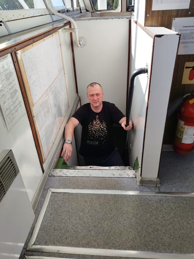 A middle-aged man with short, graying hair and a slight smile is standing in a narrow passageway, descending stairs. He is wearing a black t-shirt with a colorful, abstract design that appears to depict buildings and the word "MOSCOW" in small print below. He is holding onto a black handrail on his right side and his left hand is resting on the top step of the stairs. He is wearing a wristwatch on his left wrist.
The passageway is inside a boat or ship, indicated by the metal grate on the stairs, the textured flooring, and the presence of a fire extinguisher visible in the background. The walls are white and there are several charts or diagrams displayed on the left wall, framed by wood. Some of these charts appear to have text and drawings. A ventilation grate is visible on the far left. The lighting suggests it is daytime, with sunlight filtering in from unseen windows. The overall atmosphere is utilitarian and functional, typical of a vessel's interior. The location context is Nova, Russia.