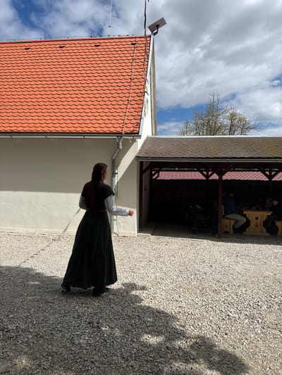 A full shot, taken outdoors, shows a woman standing beside a building in what appears to be Častá, Slovakia. The woman faces the building, with her right arm raised. She is wearing a long, dark green dress and a white blouse under a vest. She has long, dark hair. To her left is a building with a red tile roof and a white wall. Next to that building is a sheltered area with people sitting inside. The ground is covered in gravel. The sky is visible above the buildings and is partly cloudy.