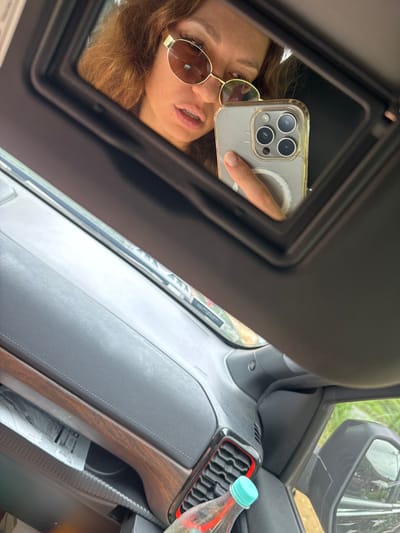 The scene shows the interior of a vehicle, likely a car, with a woman taking a selfie in the vanity mirror of the sun visor. The woman has curly hair and is wearing sunglasses. She is holding a smartphone with three camera lenses and a clear case. The car's dashboard is visible, with a water bottle in a holder near the air vents. Part of the car's side mirror is also visible. The location of Yên Minh Commune, Vietnam cannot be determined from this image.