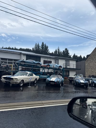 This image depicts an outdoor display of classic vehicles at what appears to be the Musée du Circuit de Spa-Francorchamps in Stavelot, Belgium, under an overcast sky with evidence of recent rain.
The scene foregrounds a wet, paved road with visible reflections. In the lower right, a black vehicle side-view mirror partially obscures the view, reflecting the wet road and distant traffic.
The main display area, also a wet paved surface, features a collection of vintage Ford-affiliated performance vehicles. A large blue multi-level car transporter truck, prominently displaying "FORD" on its side, occupies the left-center. On its upper deck, two light blue Ford GT40-style race cars are loaded; the car on the right has a white circle with the number "6". At least one additional car is visible on the transporter's lower deck.
On the ground, from left to right: a dark vintage motorcycle is parked. Next, a cream-colored 1965-66 Ford Mustang Shelby GT350 with blue racing stripes is angled towards the right, with a small informational display stand in front of it. To its right, a light blue Shelby Cobra roadster with white racing stripes is parked, angled slightly left. Further right and in the background, a white Ford F-Series pickup truck is parked, followed by two more light blue Shelby Cobra roadsters with white racing stripes.
The background consists of a two-story white painted building with a flat roof and a ground-level garage door on the left, connected to a stone-faced building on the right. A dense coniferous forest rises behind the buildings. Multiple dark power lines stretch across the upper portion of the frame. No individuals are visible, and all vehicles are static.