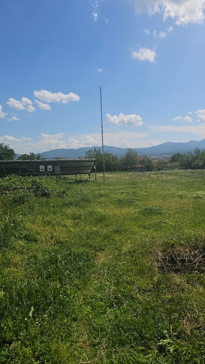 The image depicts an outdoor scene on a sunny day in Pevtsite, Bulgaria. The sky is blue with scattered white clouds. A tall metal pole extends vertically into the sky. In the foreground, there is a field of green grass. Behind the grass, there are solar panels. Behind the solar panels are trees and a mountain range.