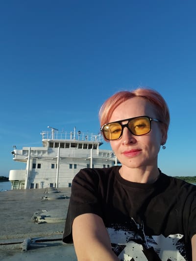 A woman with short, pink-tinted hair and yellow-tinted sunglasses is taking a selfie outdoors under a clear blue sky. She is wearing a black t-shirt with a graphic design. In the background, a large, white ship with multiple decks and windows is docked. The foreground shows a weathered, grey surface with various metal fixtures. The image appears to be taken on a sunny day. The scene is located in Kochino, Russia.