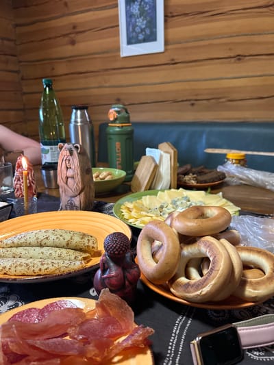 The image captures a table setting with a variety of food items, suggesting a meal or gathering. The food includes bagels, cheeses, pancakes, and sliced fish. A wooden carving and a candle are also present on the table. A thermos, a bottle of olive oil, and a framed picture are visible in the background. The location is Лесные Поляны, Russia.