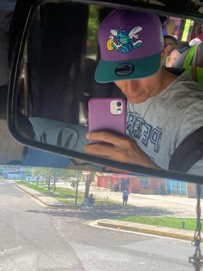The image captures a selfie in a vehicle mirror, showing a person wearing a grey t-shirt, a purple and green Charlotte Hornets baseball cap, and a silver chain, holding a purple phone. The reflection reveals a street in San José de Barlovento, Venezuela, with buildings, trees, sidewalks, and people walking.
