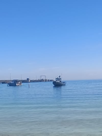 In Oakville, Canada, a bright, clear blue sky dominates the upper portion of the image, with a thin haze near the horizon. The lower two-thirds of the image depict a calm, blue body of water. In the mid-ground, a long, concrete pier extends across the scene, supported by pilings and featuring various structures, including lights and what appears to be a gantry or elevated walkway.

Two boats are visible on the water. To the left, a sleek, white motorboat carries several individuals who are seated and appear to be relaxing. It is positioned near a light-blue inflatable mat. To the right, a dark-colored rigid inflatable boat (RIB) is underway, with a single person visible in the cockpit steering.

In the foreground, the water transitions to a lighter, more translucent blue, revealing a sandy or pebbly bottom beneath the surface. The overall scene conveys a peaceful, recreational atmosphere on a sunny day.