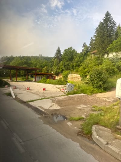 This image depicts a neglected roadside environment in Boichovtsi, Bulgaria. The scene is viewed from a slightly elevated position, with motion blur evident on the road in the immediate foreground, suggesting capture from a moving vehicle.

The foreground on the left features a dark asphalt road with faint white markings and a metal grate embedded along its edge. To the right of the road, a cracked concrete and dirt area is visible, containing several shallow puddles of water. This area is partially defined by a low concrete curb.

The midground is dominated by what appears to be an abandoned gas station. Two overhead canopy structures, supported by dark red/maroon painted steel beams, stand over a heavily cracked and weed-infested concrete forecourt. The larger canopy is positioned towards the left, while a smaller one is further back and to the right. Beneath the larger canopy, concrete foundations for what were likely fuel pumps are visible. Behind the smaller canopy, a small, single-story building with yellow trim and windows, presumably the former service station, is discernible. Multiple white utility poles or streetlights are scattered throughout the station area. Several red and white striped concrete blocks act as barriers or markers on the paved surface. A grey wheeled trash bin is located on the right side of the forecourt, and a white portable toilet is partially visible on the far right edge of the frame.

The background consists of a dense forest covering a rising hillside, featuring a mix of deciduous and coniferous trees. A concrete or stone retaining wall, partially covered by vegetation, is visible behind the station structures. Further up the hill and integrated into the tree line, a small building with a reddish-brown roof and light-colored walls is partially visible. The sky overhead shows patches of light blue with dispersed white clouds, some appearing wispy and others more substantial, with a general haziness in the upper left quadrant. No people are present in the image.