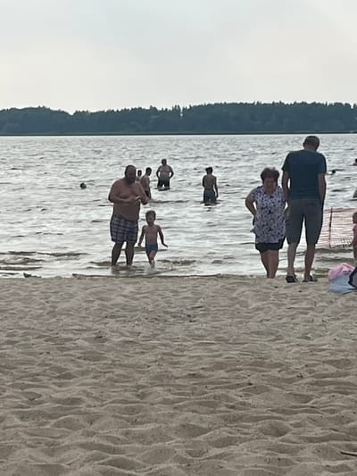 The image shows a group of people at a beach, with the water and sand in the foreground. The setting appears to be a lake or calm body of water with a tree-lined shore in the background. The time of day seems to be overcast, possibly late afternoon or evening, as the light is muted.
In the water, several individuals are visible. Towards the left, a man in plaid shorts is holding a child, and another child is standing nearby. Further back, there are other individuals in the water, some appearing to be adults and others possibly children. On the sandy shore, a man in a dark t-shirt and shorts stands next to a woman wearing a patterned top and shorts. A pink bag and a white object, possibly another bag or clothing item, are also on the sand behind them.
The people seem relaxed and engaged in typical beach activities, such as wading in the water or relaxing on the shore. The overall mood appears to be casual and recreational. There is no visible text in the image. The location context is described as Загародскі сельскі Савет, Belarus.